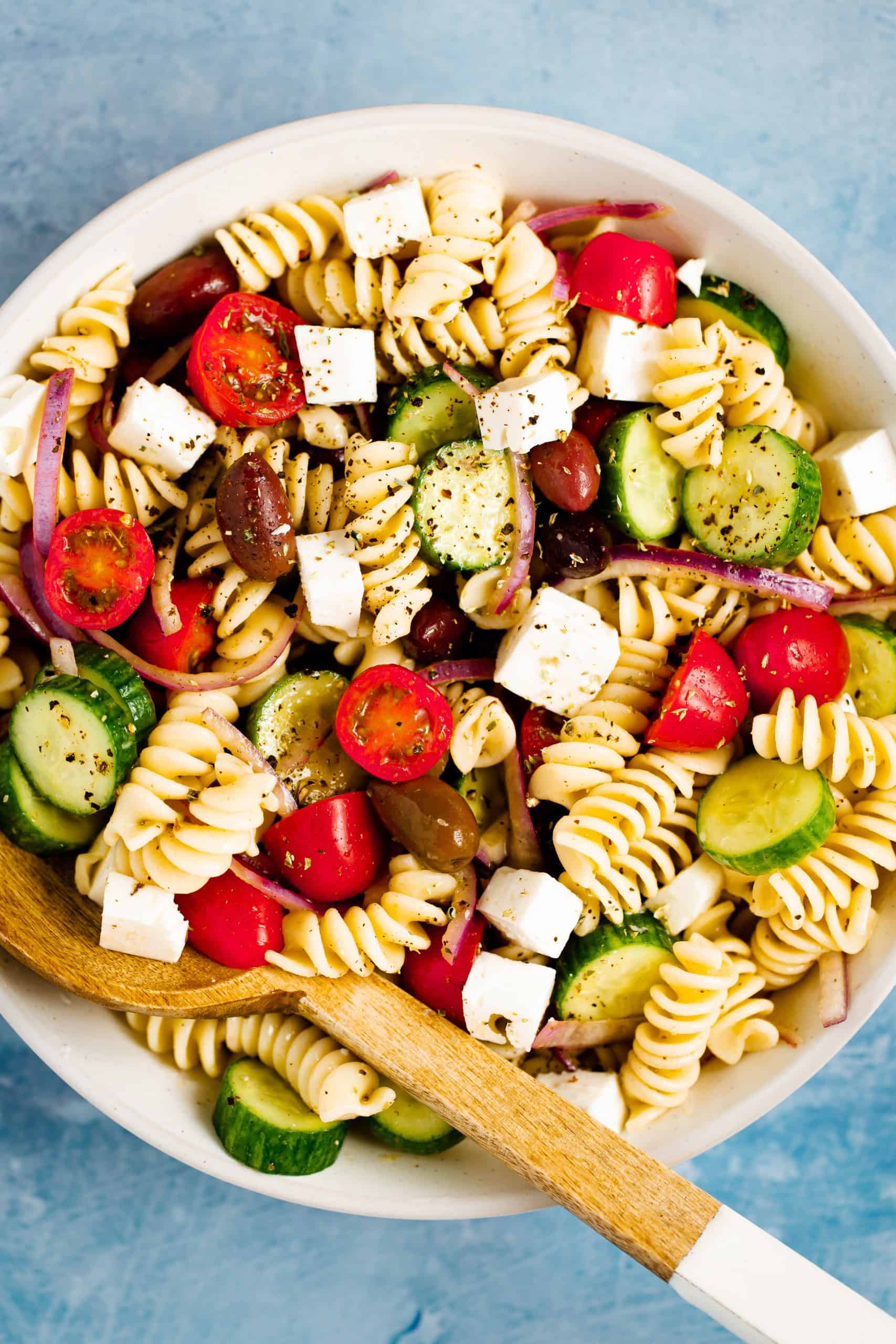 White bowl filled with greek pasta salad and a wooden salad spoon
