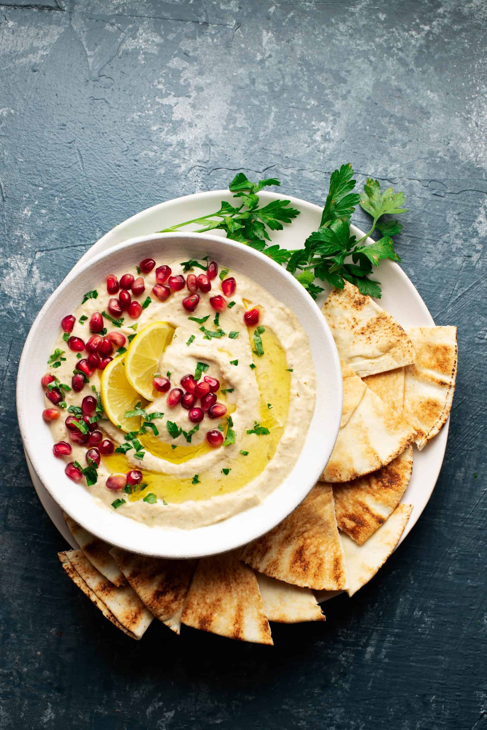 White bowl of Baba ganoush topped with parsley, olive oil, and pomegrante on a white plate with pita bread. 