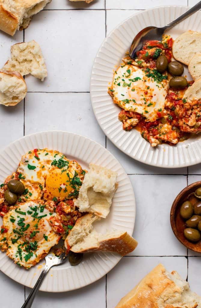 two plates of shakshuka and ripped bread with a wooden plate of green olives