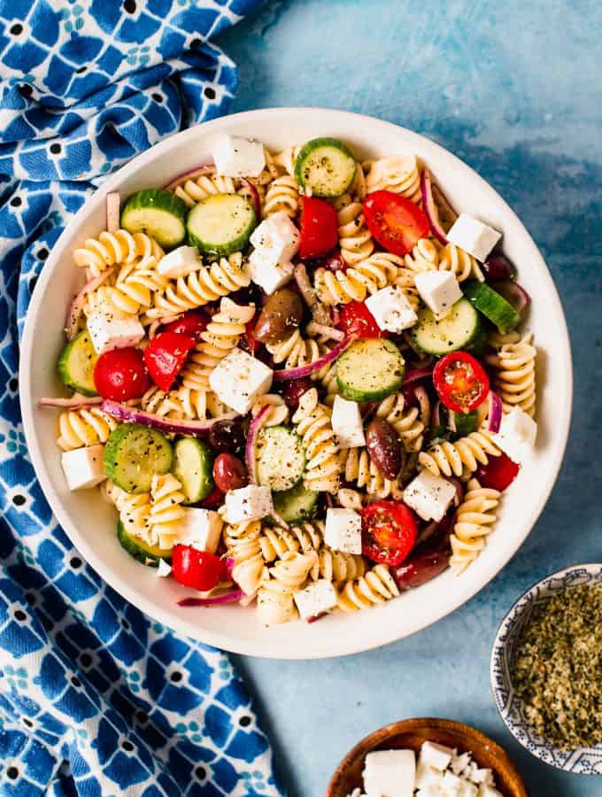 Pasta salad in a white bowl with small bowl of oregano and small bowl of feta cheese