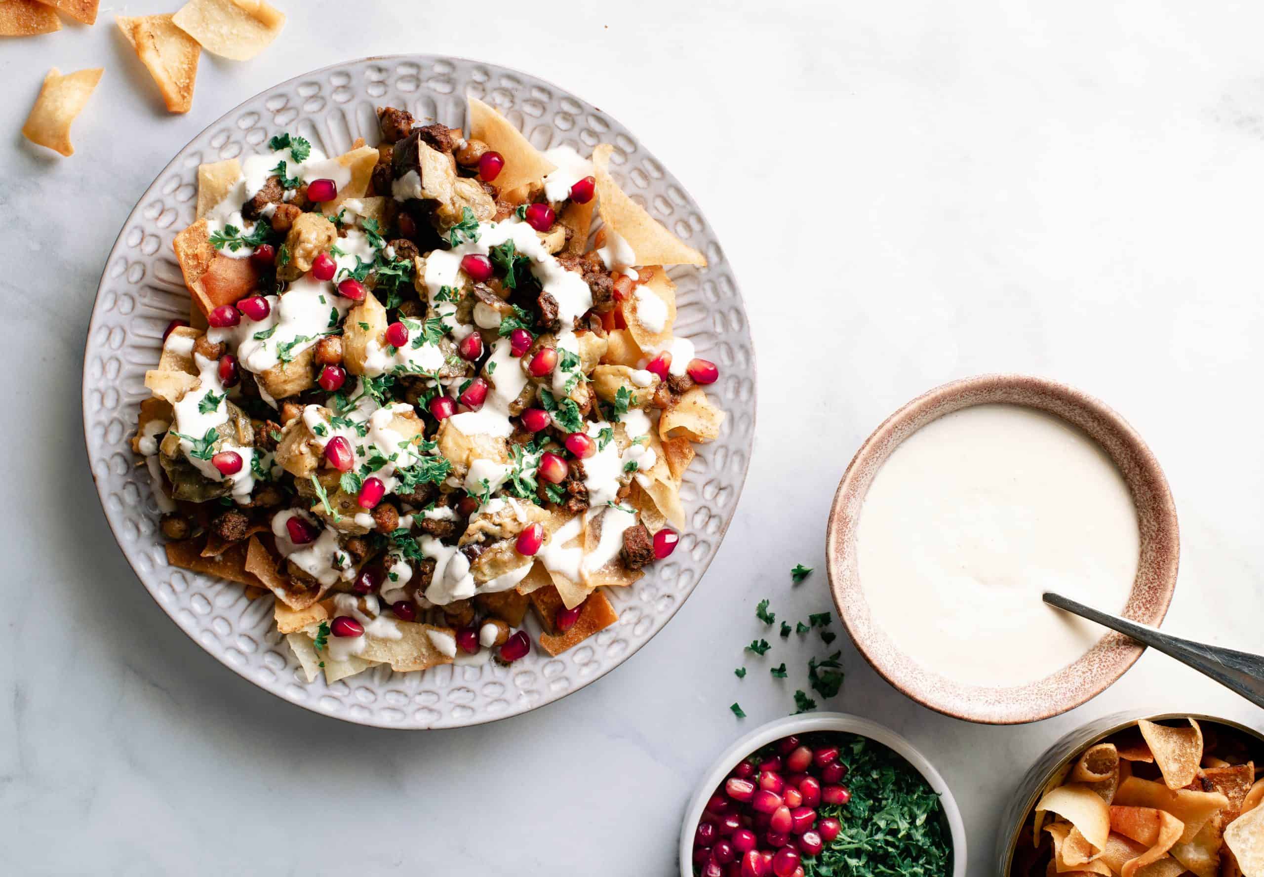 A plate full of Eggplant & Beef Fettah with small bowls of the ingredient: yogurt sauce, pita chips, parsley, & pomegrantes