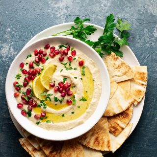 White bowl of pureed eggplant topped with parsley, olive oile, and pomegrante on a white plate with pita bread.