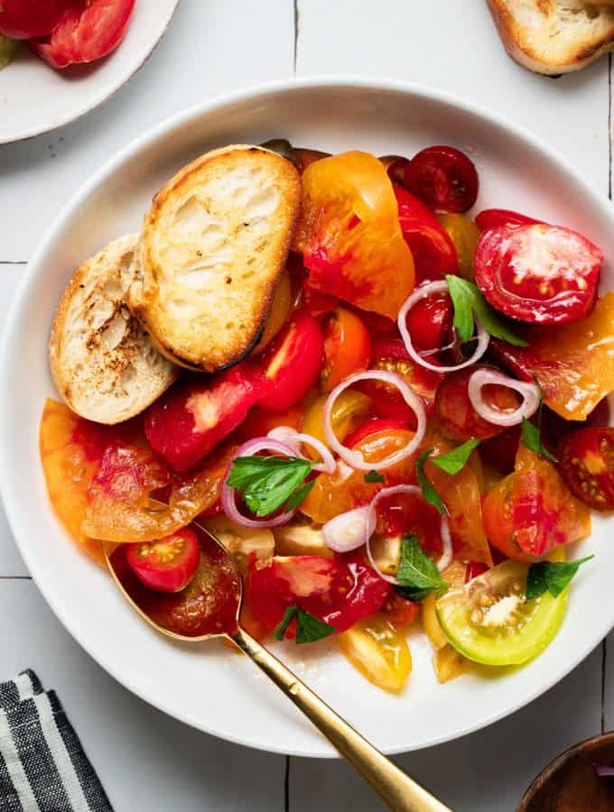 a white bowl filled with heirloom tomatoes, a golden serving spoon and a few pieces of bread